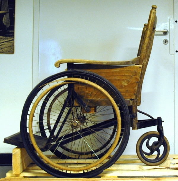 Rullstol i trä med tre hjul, två stora och ett mindre. Märkt på baksidan "KSA6an".