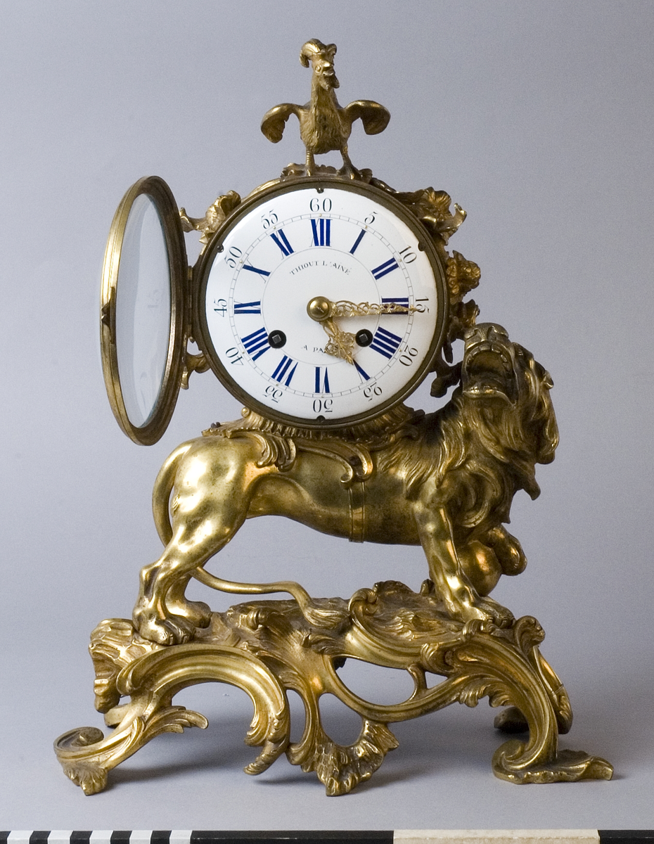 Urhuset i form av en tunna som står på ett rytande lejon med ena foten på ett klot på ett genombrutet rocailleornament. Ovanpå tunnan står en galande tupp, på sidorna rosor och växter.