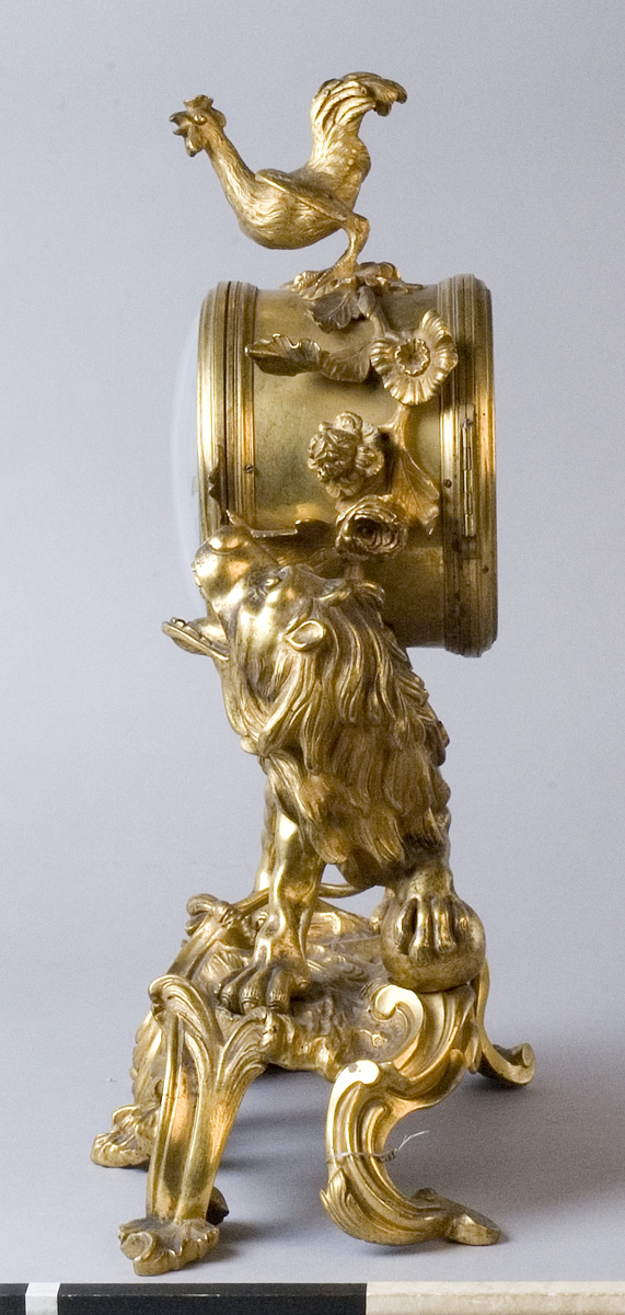 Urhuset i form av en tunna som står på ett rytande lejon med ena foten på ett klot på ett genombrutet rocailleornament. Ovanpå tunnan står en galande tupp, på sidorna rosor och växter.