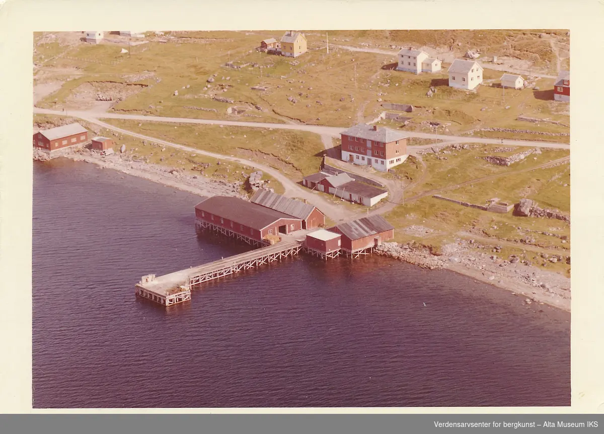 Oversiktsbilde fra Widerøe av Bossekop med utsikt over Alta fjordhotell, flere bolighus, kai, sjøhus og veien som går mellom dem. 