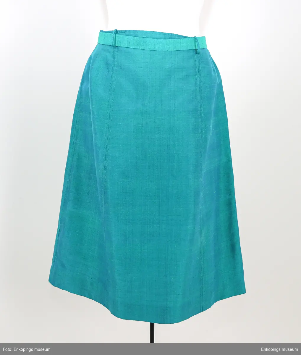 Turkosgrön med blått foder är kjolen och är sydd i sex våder, saknas linning men har ett skärp i tyget. Väska EM7293 är ämnad att bäras till denna dräkt. Enligt givaren en sömmerska i Norrland.