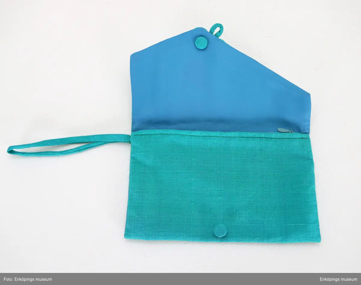 Turkosgrönt kuvert med hank som handtag och konsfullt gjord knäppning. Har blått foder och dragkedja inuti. Ämnad att användas tills. med EM7294.