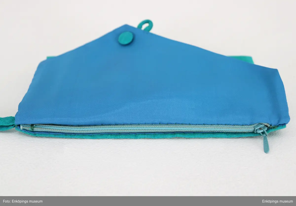 Turkosgrönt kuvert med hank som handtag och konsfullt gjord knäppning. Har blått foder och dragkedja inuti. Ämnad att användas tills. med EM7294.