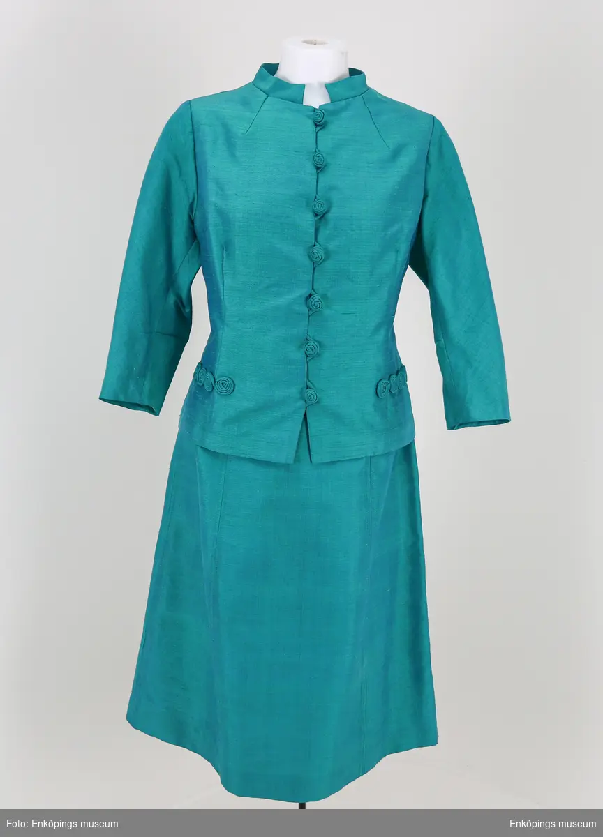 Turkosgrön med blått foder är kjolen och är sydd i sex våder, saknas linning men har ett skärp i tyget. Väska EM7293 är ämnad att bäras till denna dräkt. Enligt givaren en sömmerska i Norrland.