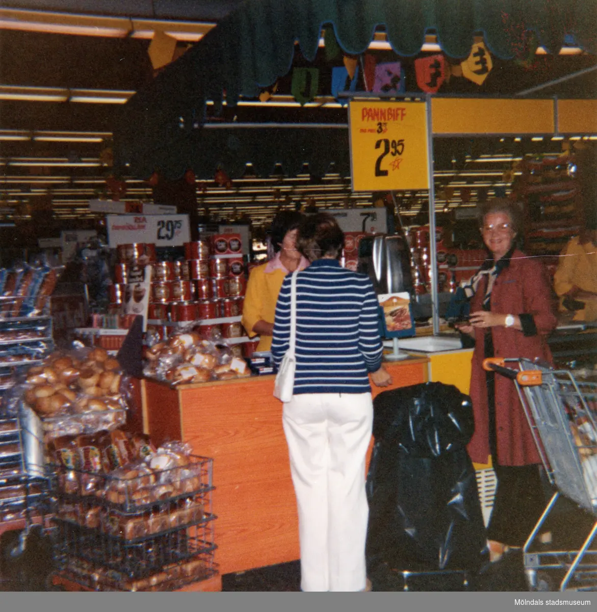 En kvinnlig demonstratris, klädd i gul rock, står vid en disk för provsmakning av bröd, Domus Mölndal C efter 1976. En kund och en expedit står framför disken. I bakgrunden ses stora, runda, uppstaplade aluminiumburkar med Cirkelkaffe.