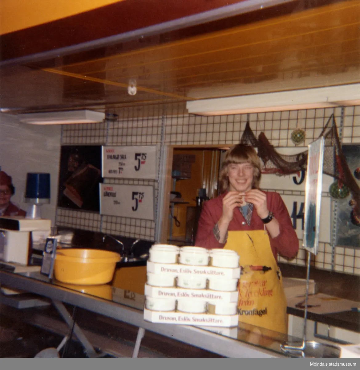 En kvinnlig expedit, klädd i röd rock och gult förkläde, står bakom fiskdisken hållandes en liten räka i händerna, Domus Mölndal C efter 1976. På disken ses förpackningar från Druvan, Eslöv, Smaksättare. På den vit-kaklade väggen i bakgrunden ses skyltar med olika sill-erbjudanden från Winner (Konsum-produkt).