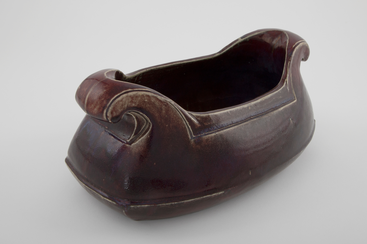 Støpt steingods med rødlilla feltspattgalsur med enkelte blå felt. Avlangt jardinière med to håndtak. Håndtakene ruller seg utover og nedover mot korpus. Linjer langs håndtak og åpning som understreker formen. Fordypet linje over mellom base og korpus.