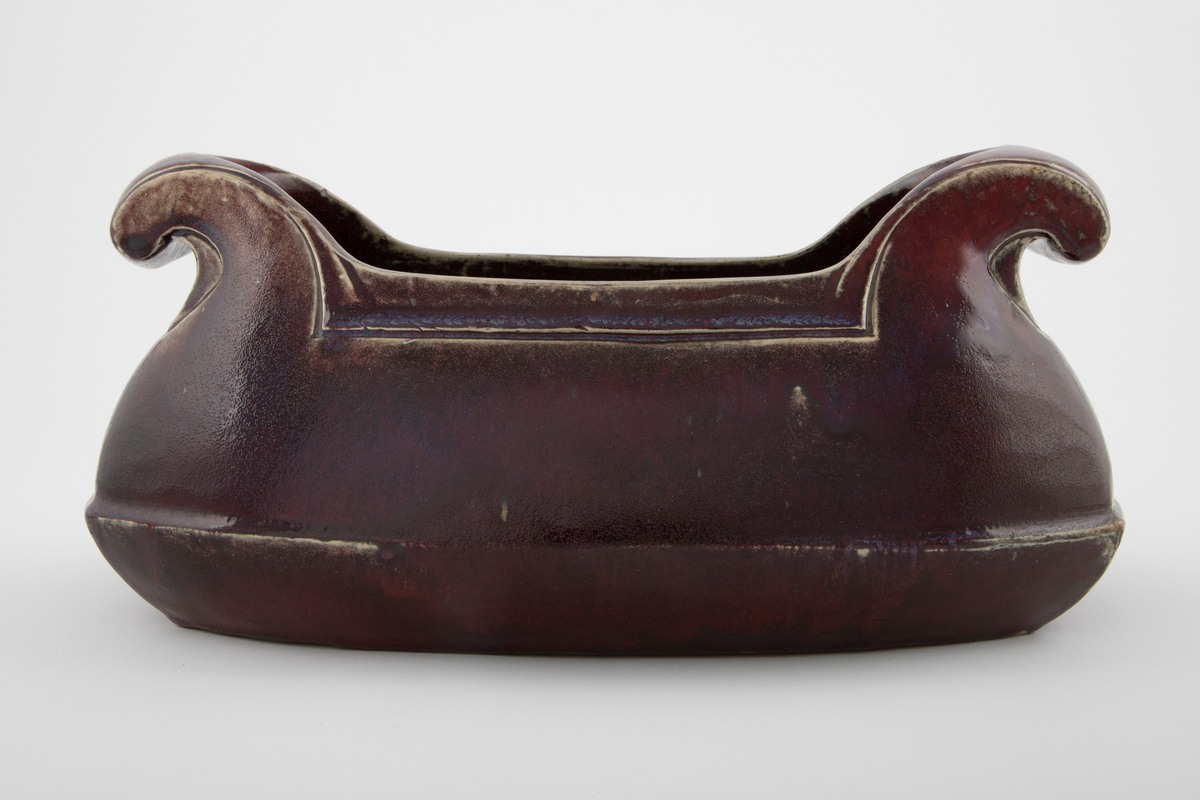 Støpt steingods med rødlilla feltspattgalsur med enkelte blå felt. Avlangt jardinière med to håndtak. Håndtakene ruller seg utover og nedover mot korpus. Linjer langs håndtak og åpning som understreker formen. Fordypet linje over mellom base og korpus.