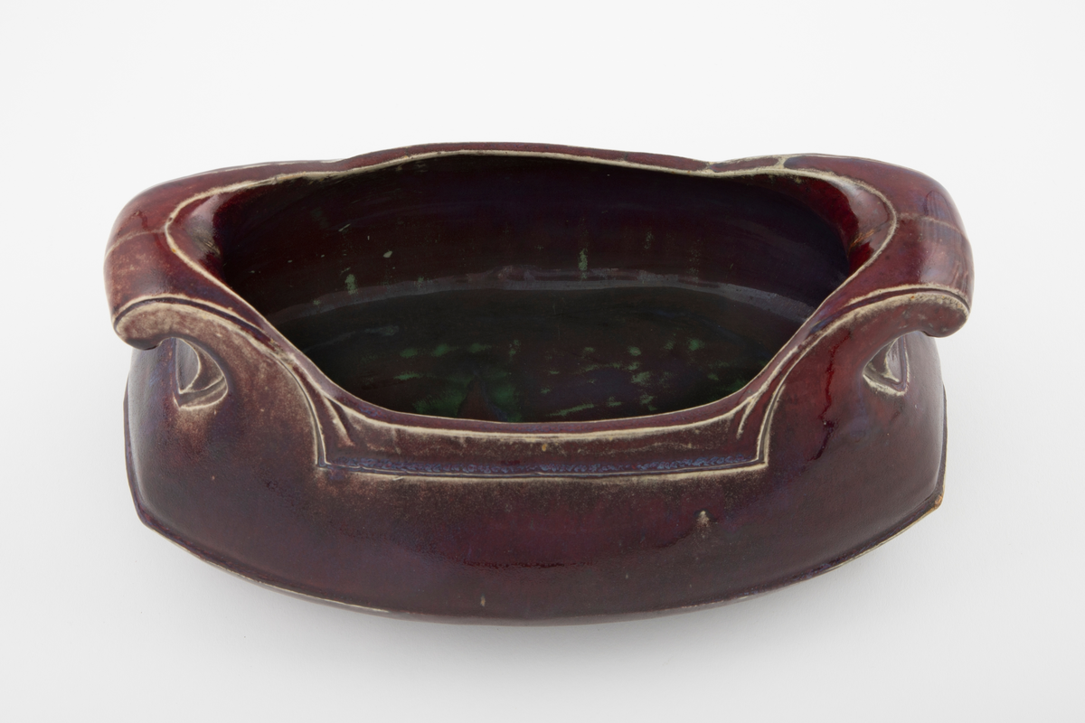 Støpt steingods med rødlilla feltspattgalsur med enkelte blå felt. Avlangt jardinière med to håndtak. Håndtakene ruller seg utover og nedover mot korpus. Linjer langs håndtak og åpning som understreker formen. Fordypet linje over mellom base og korpus.