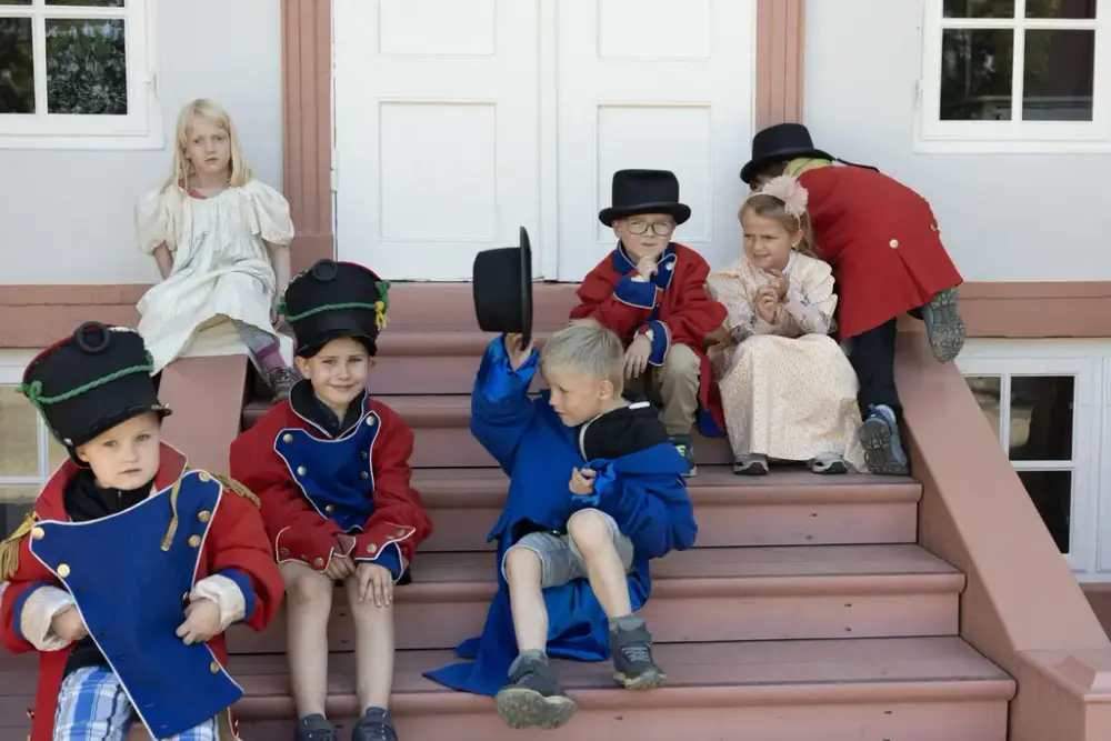 Barn kledd i historiske kostymer sitter på trappen foran Eidsvollsbygningen.