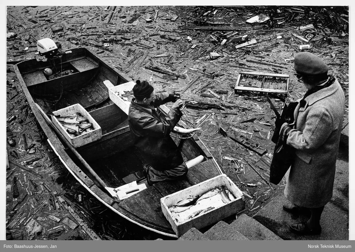 En kvinne kjøper fisk av en fisker i en jolle ved Rådhuskaia i Oslo. Vannet rundt jolla er fylt med trevirke og annet søppel. 