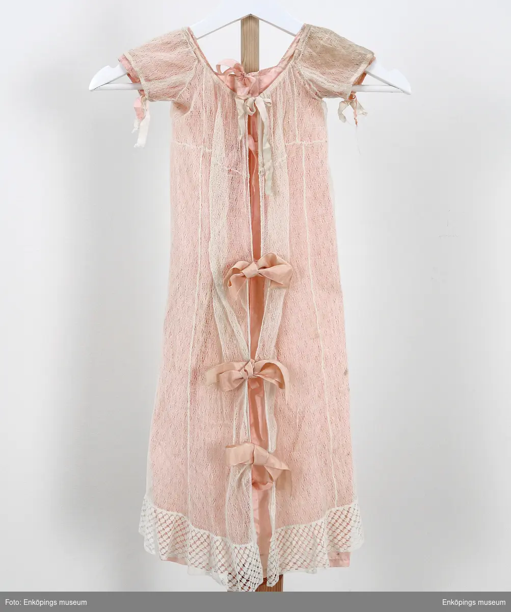 Rosa sidenunderkjol samt vit spetsklänning med knypplad volang. Sidenband att knyta i ryggen. Både använd och tillverkad på ca 1840 talet. Har nog förmodligen blivit buren av pastor Spaks tre fastrar.