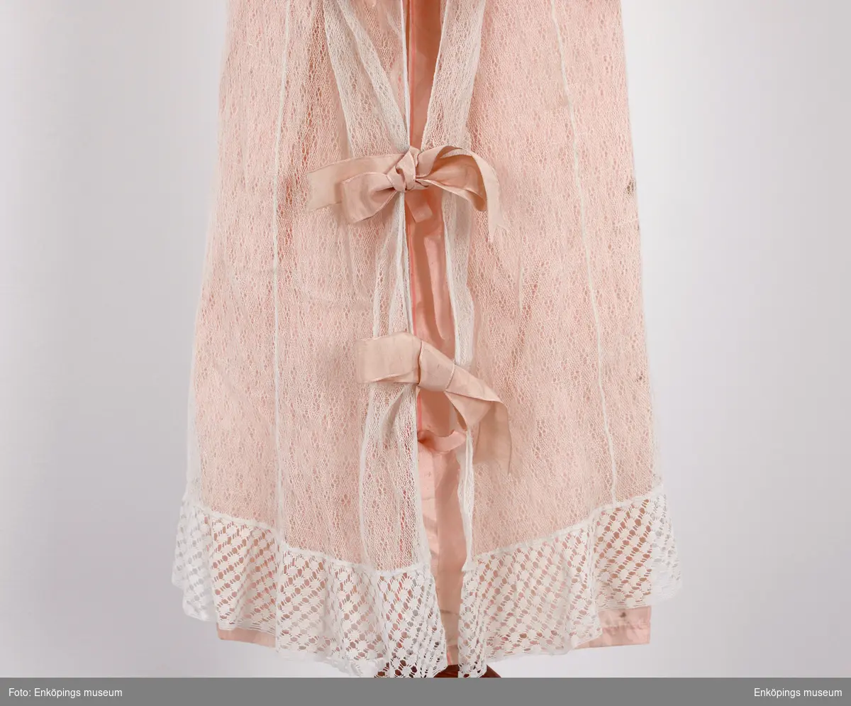 Rosa sidenunderkjol samt vit spetsklänning med knypplad volang. Sidenband att knyta i ryggen. Både använd och tillverkad på ca 1840 talet. Har nog förmodligen blivit buren av pastor Spaks tre fastrar.