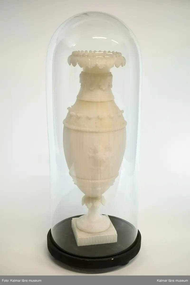 KLM 17915:1. Urna, prydnadsurna. Av alabaster och med tillhörande glaskupa. Formad som en vas på fot med fyrkantig sockel. Med skulpterad dekor i form av flera akantusbårder, pärlstav, samt med centrerad blomsterbukett i fronten. Mittstycke och sockel med kannelyr. Förvaras i glaskupa med bottenplatta av trä i svart färg. Urnan delbar i två delar. Hör samman med KLM 17915:2 Urna.