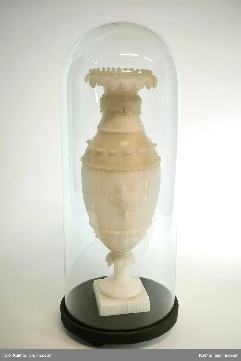 KLM 17915:2. Urna, prydnadsurna. Av alabaster och med tillhörande glaskupa. Formad som en vas på fot med fyrkantig sockel. Med skulpterad dekor i form av flera akantusbårder, pärlstav, samt med centrerad blomsterbukett i fronten. Mittstycke och sockel med kannelyr. Förvaras i glaskupa med bottenplatta av trä i svart färg. Urnan delbar i tre delar, samt med sprickor och skadad dekor. Hör samman med KLM 17915:1 Urna.