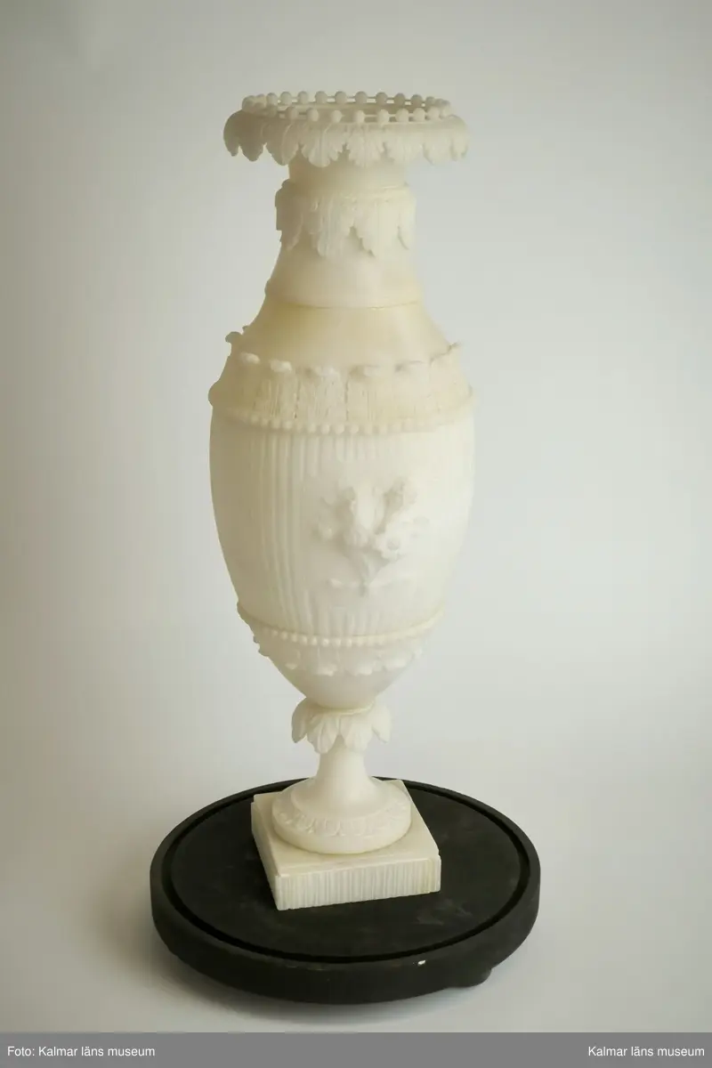 KLM 17915:1. Urna, prydnadsurna. Av alabaster och med tillhörande glaskupa. Formad som en vas på fot med fyrkantig sockel. Med skulpterad dekor i form av flera akantusbårder, pärlstav, samt med centrerad blomsterbukett i fronten. Mittstycke och sockel med kannelyr. Förvaras i glaskupa med bottenplatta av trä i svart färg. Urnan delbar i två delar. Hör samman med KLM 17915:2 Urna.
