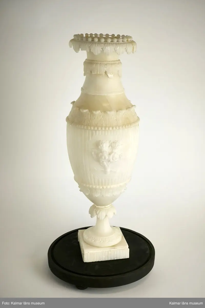 KLM 17915:2. Urna, prydnadsurna. Av alabaster och med tillhörande glaskupa. Formad som en vas på fot med fyrkantig sockel. Med skulpterad dekor i form av flera akantusbårder, pärlstav, samt med centrerad blomsterbukett i fronten. Mittstycke och sockel med kannelyr. Förvaras i glaskupa med bottenplatta av trä i svart färg. Urnan delbar i tre delar, samt med sprickor och skadad dekor. Hör samman med KLM 17915:1 Urna.