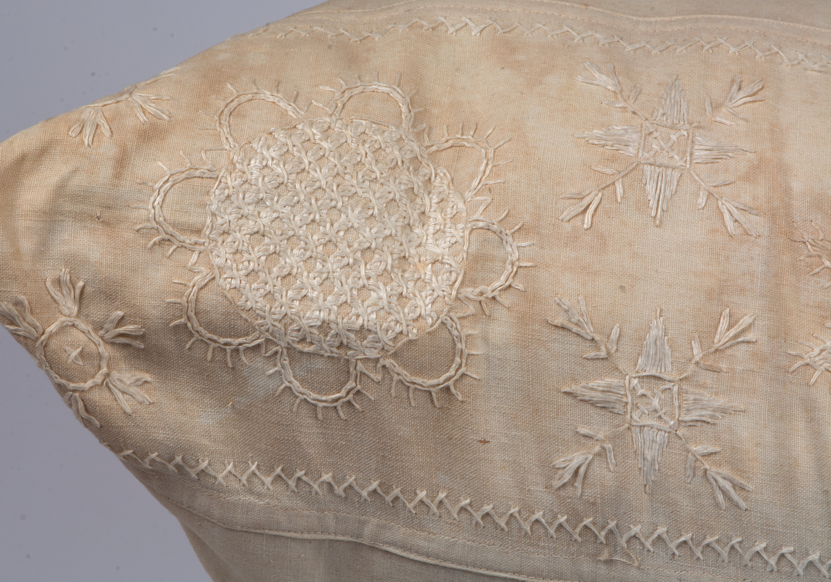 Kuddvar av vitt linnetyg med broderad bård på en kortsida, hallandssöm. Tyget är mycket gulnat och har några hål. Tygklädda knappar i ena kortsidan. Hålsöm vid linning. Sitter på kudde VMF100321.
