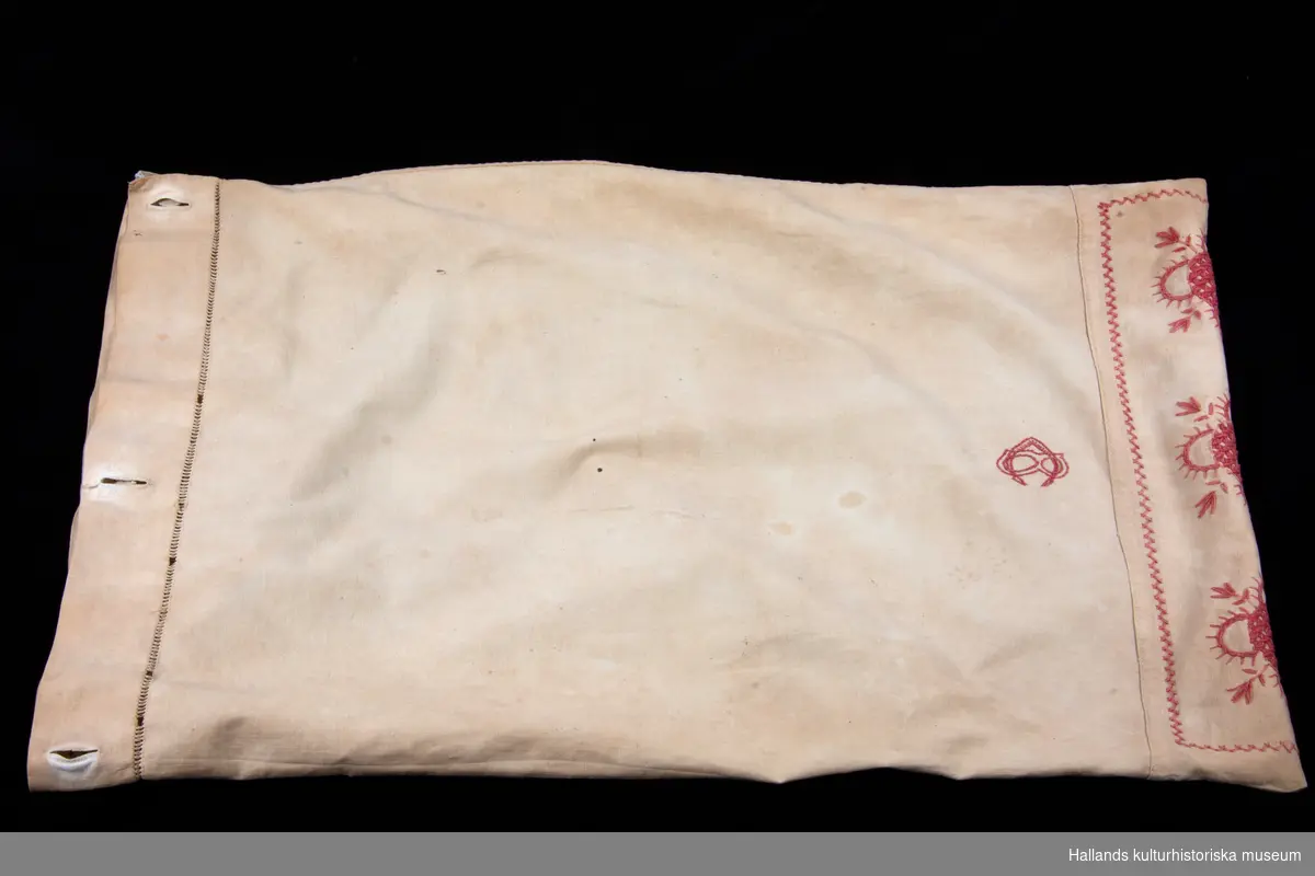 Örngott av vävt bomullstyg med klädda knappar i öppningen. Kanten utmed öppningen ör sydd med hålsöm. Ena sidan broderad med röd hallandssöm  Broderade initialer: "CB" i jugendstil.