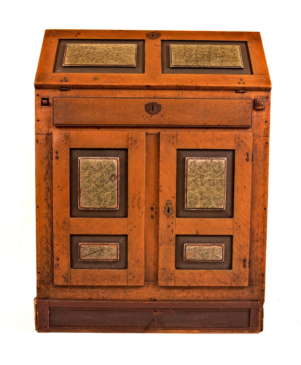 Skatholl med skuff over og under dobbel dør. Inndela med 8 små skuffar og eit ope rom bak skriveklaff. Lås og nykil til dør og til skriveklaff. Ådringsmåla.