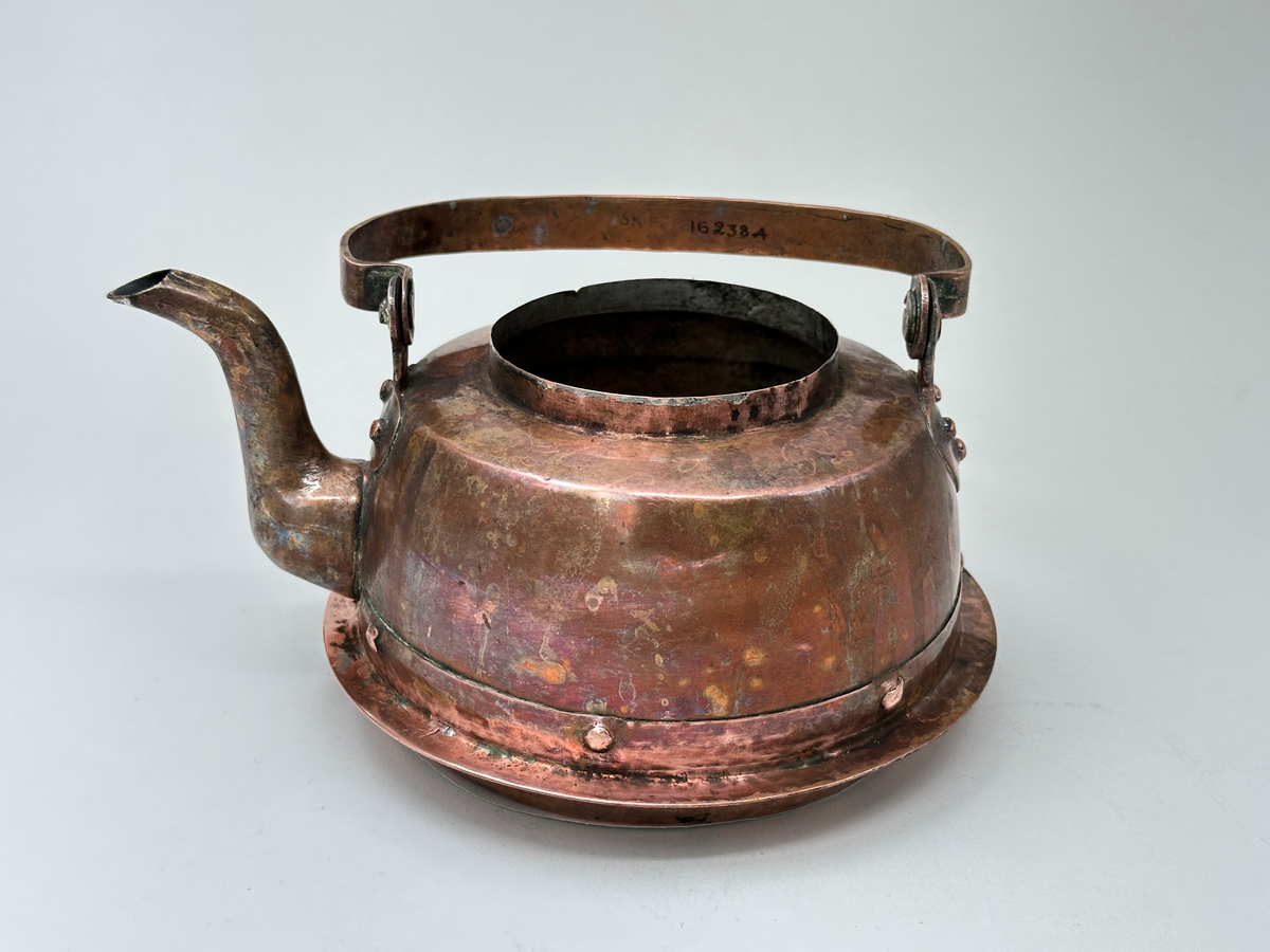 Kaffepanna, driven av koppar, med insidan förtent. Modell med plattrund form, fällbart bygelhandtag, svängd pip. Ena bygelfästet avlångt, andra hjärtformat. Synlig skarv med flikar och mässingsslaglod. Pannan har en fastnitad fals, avsedd för järnspis. 