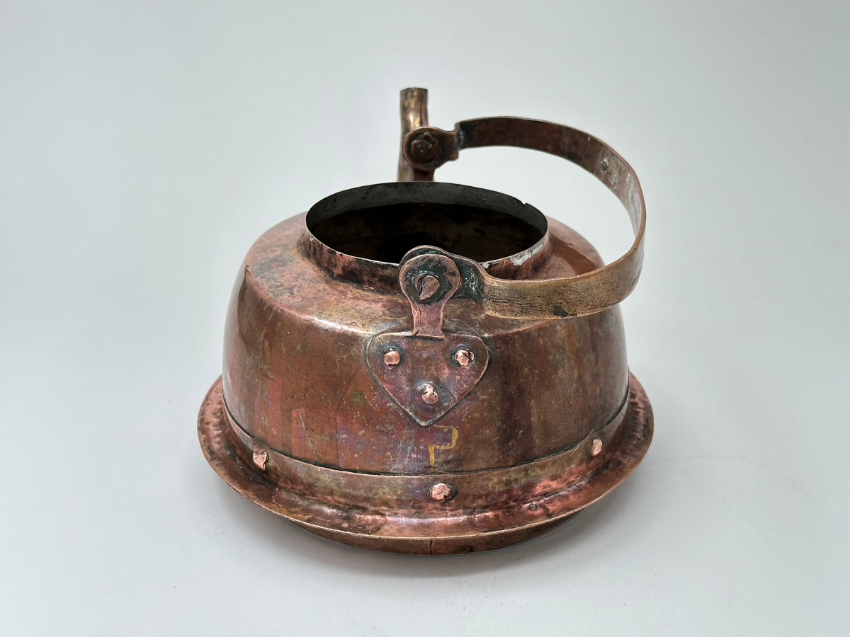 Kaffepanna, driven av koppar, med insidan förtent. Modell med plattrund form, fällbart bygelhandtag, svängd pip. Ena bygelfästet avlångt, andra hjärtformat. Synlig skarv med flikar och mässingsslaglod. Pannan har en fastnitad fals, avsedd för järnspis. 