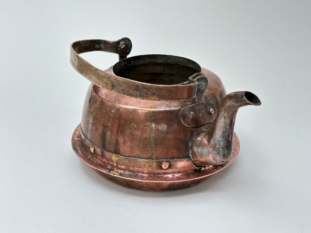 Kaffepanna, driven av koppar, med insidan förtent. Modell med plattrund form, fällbart bygelhandtag, svängd pip. Ena bygelfästet avlångt, andra hjärtformat. Synlig skarv med flikar och mässingsslaglod. Pannan har en fastnitad fals, avsedd för järnspis. 