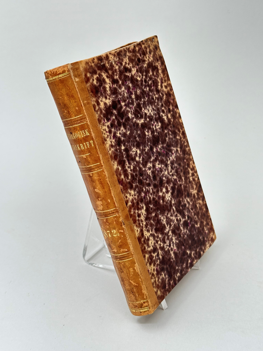 Bok: "Pedagogisk tidskrift 1872". Tryckt 1872 i Stockholm.
Halvfranskt band. Rygg av cognacsfärgat läder med förgyllda nedsänkta bokstäver och rader. Hörn förstärkta med läder. Pärm av kartong klädd i fläckmarmorerat papper i brunt och gult.