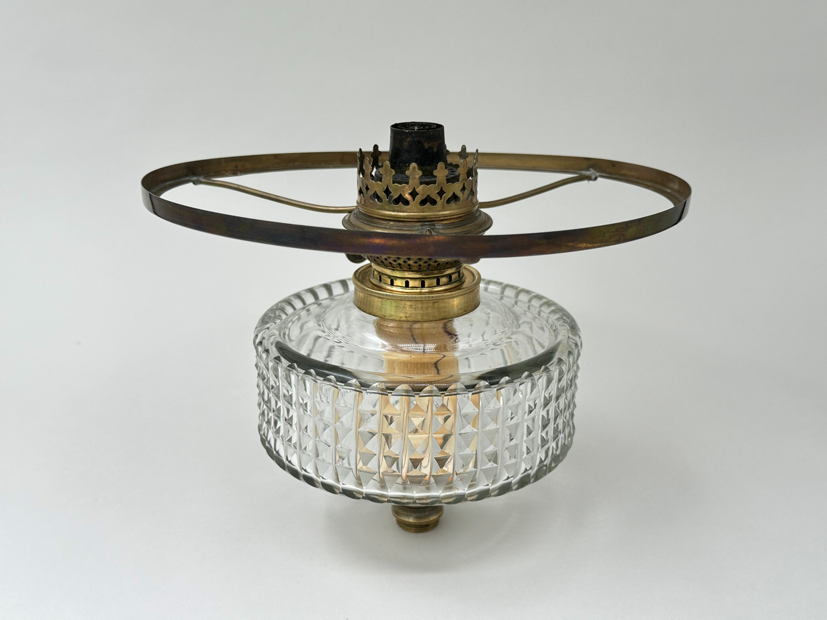 Oljehus och brännare till fotogenlampa. Oljehus i klart glas, slipat i diamantslipning som övergår till olivslipning i över- och nederkant. Dekorativt utformad brännare i mässing med påkopplad ställning för att hålla lampskärmen. Ställningen defekt, och en av dess armar är fastlödda. 