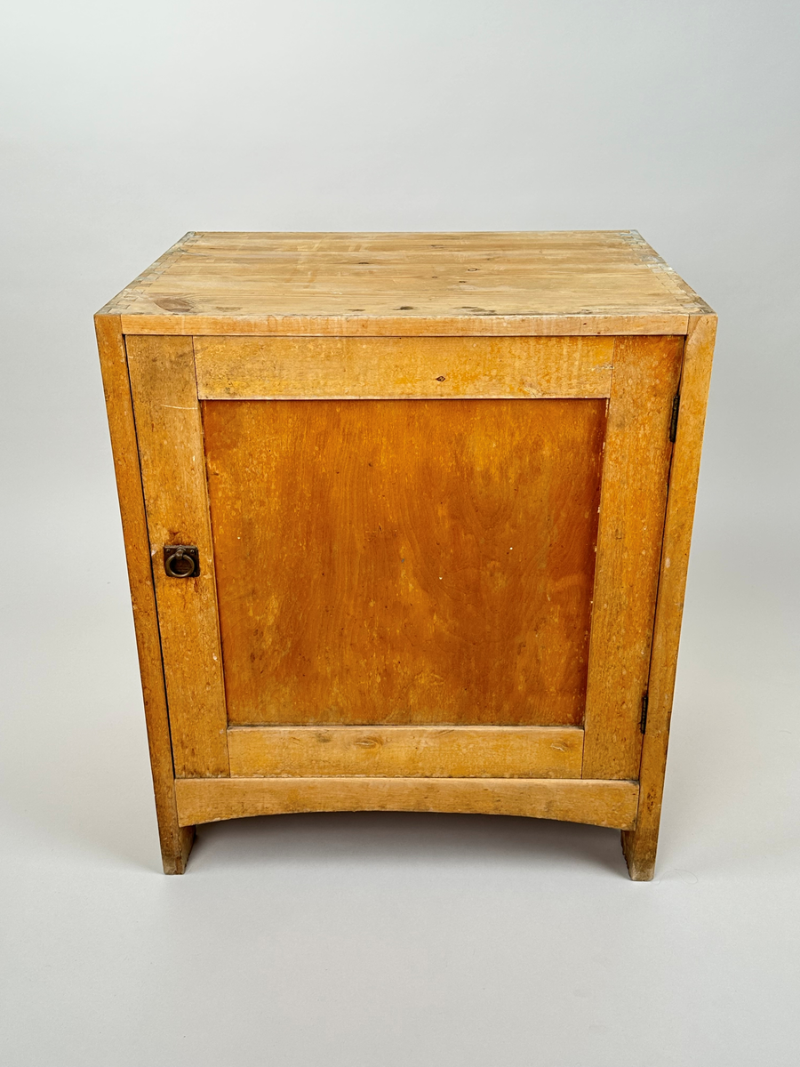 Kommod tillhörande tvättställ. I trä, furu med björkfanér. Sinkade sidor. På framsidan en utåtgående dörr med spegel och ringhandtag i metall. Gavlarna har bågformade urtag längst ner. På baksidan är blindträ i furu synligt.