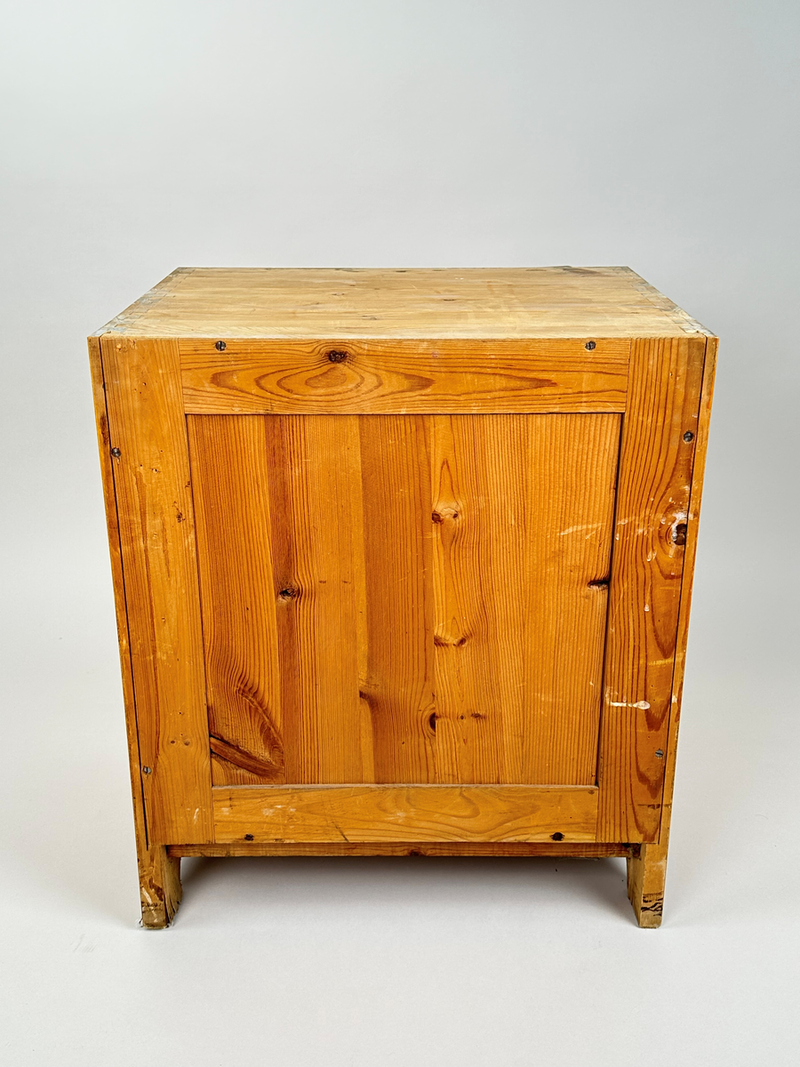 Kommod tillhörande tvättställ. I trä, furu med björkfanér. Sinkade sidor. På framsidan en utåtgående dörr med spegel och ringhandtag i metall. Gavlarna har bågformade urtag längst ner. På baksidan är blindträ i furu synligt.