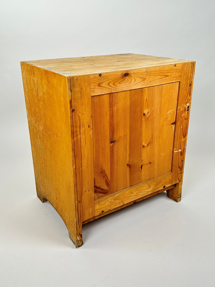 Kommod tillhörande tvättställ. I trä, furu med björkfanér. Sinkade sidor. På framsidan en utåtgående dörr med spegel och ringhandtag i metall. Gavlarna har bågformade urtag längst ner. På baksidan är blindträ i furu synligt.