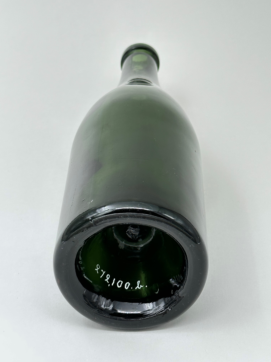 Butelj, av grönt glas. Upphöjd botten, s.k. kinnekulle. Tjock glasmassa med en hel del lodräta ojämnheter. Spår av form. 89 i relief i botten (väldigt svagt).