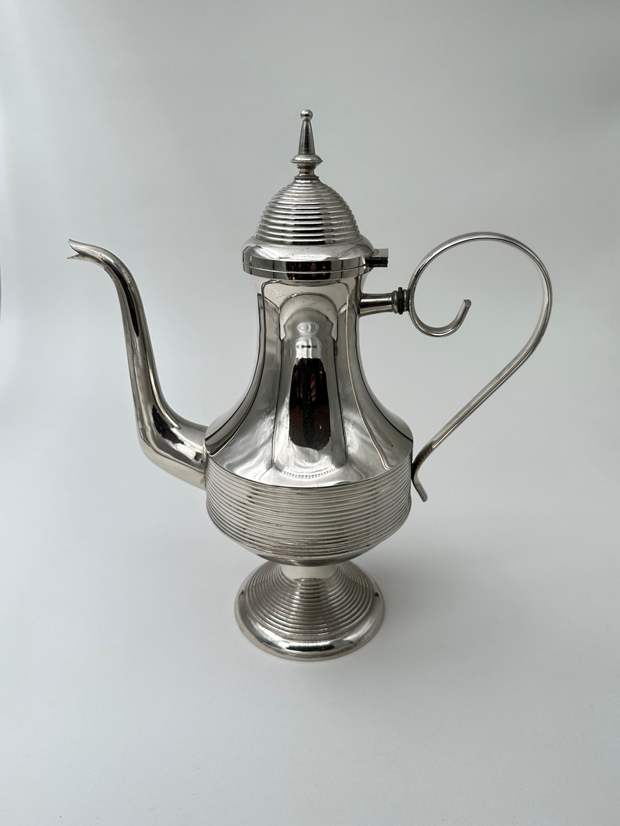 Kaffekanna i vitmetall. Välvd, vågrätt räfflad fot, avsmalnande upptill. Kroppen vägrätt räfflad nedtill och konkavt avsmalnande. Lock vågrätt räfflat, konvext avsmalnande och avslutas i en avlång lockknopp. Dekorativt formad grepe, fäst i kannans hals med ett stöd i metall. Avsmalnande slät lätt rundad pip. 