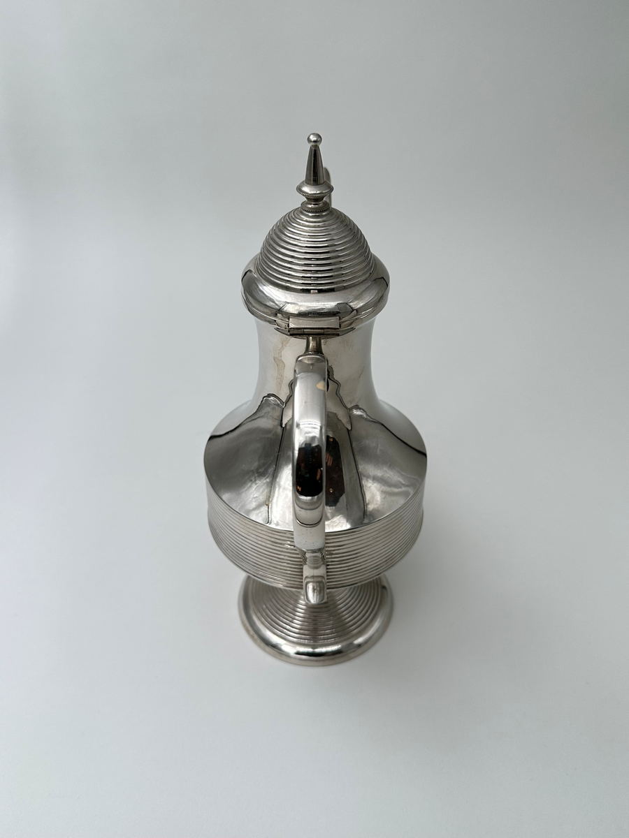 Kaffekanna i vitmetall. Välvd, vågrätt räfflad fot, avsmalnande upptill. Kroppen vägrätt räfflad nedtill och konkavt avsmalnande. Lock vågrätt räfflat, konvext avsmalnande och avslutas i en avlång lockknopp. Dekorativt formad grepe, fäst i kannans hals med ett stöd i metall. Avsmalnande slät lätt rundad pip. 