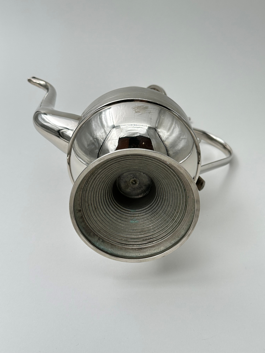 Kaffekanna i vitmetall. Välvd, vågrätt räfflad fot, avsmalnande upptill. Kroppen vägrätt räfflad nedtill och konkavt avsmalnande. Lock vågrätt räfflat, konvext avsmalnande och avslutas i en avlång lockknopp. Dekorativt formad grepe, fäst i kannans hals med ett stöd i metall. Avsmalnande slät lätt rundad pip. 