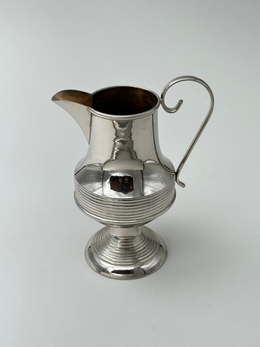 Gräddkanna i vitmetall. Välvd, vågrätt räfflad fot, avsmalnande upptill. Kroppen vägrätt räfflad nedtill och avsmalnande upptill. Mynningen övergår i kannans snip. Dekorativt formad grepe i metall. 