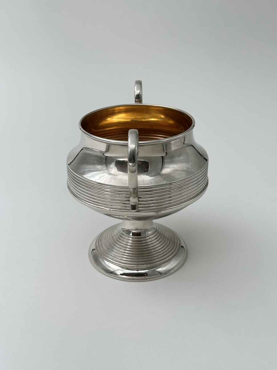 Rund sockerskål i vitmetall. Välvd, vågrätt räfflad fot, avsmalnande upptill. Kroppen vägrätt räfflad nedtill och konkavt avsmalnande. Rundad kant. Dekorativt formade grepar i metall.