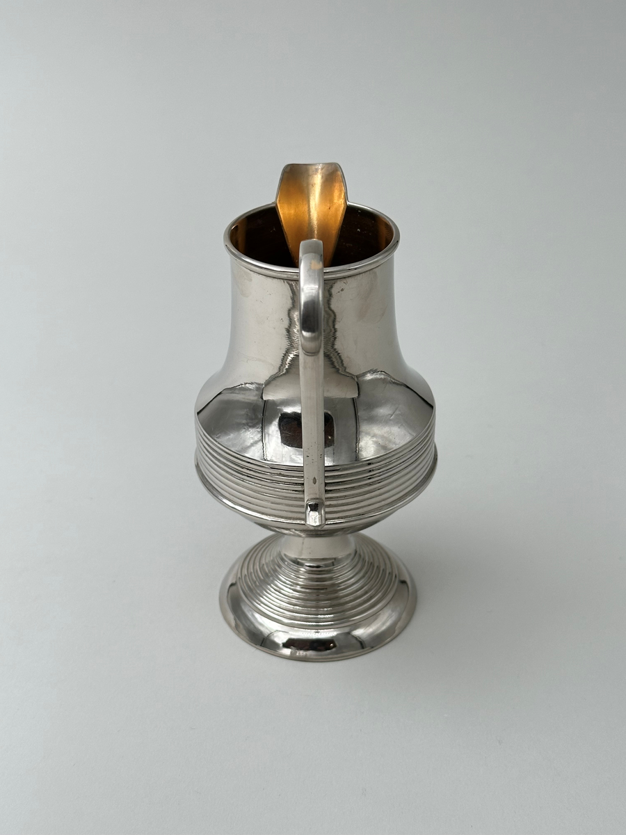 Gräddkanna i vitmetall. Välvd, vågrätt räfflad fot, avsmalnande upptill. Kroppen vägrätt räfflad nedtill och avsmalnande upptill. Mynningen övergår i kannans snip. Dekorativt formad grepe i metall. 