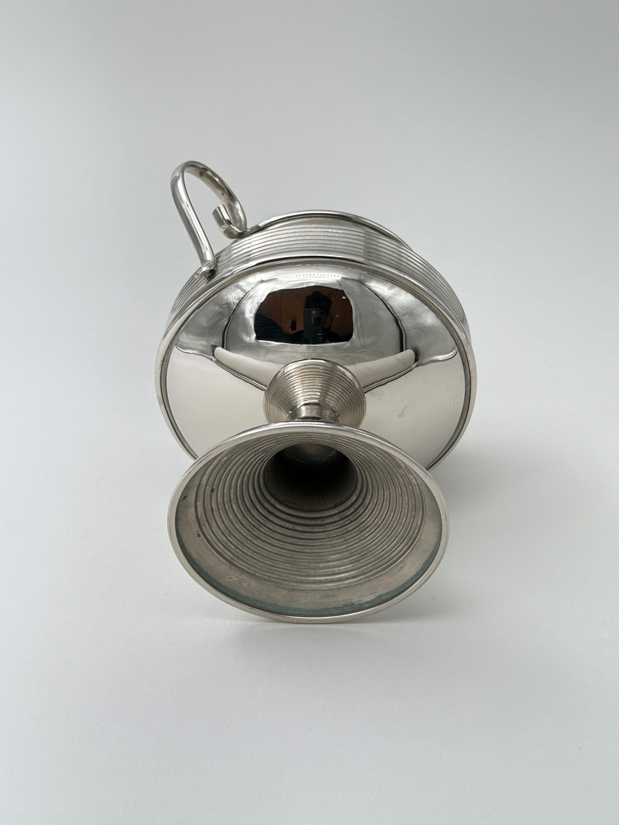 Rund sockerskål i vitmetall. Välvd, vågrätt räfflad fot, avsmalnande upptill. Kroppen vägrätt räfflad nedtill och konkavt avsmalnande. Rundad kant. Dekorativt formade grepar i metall.