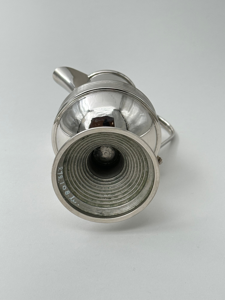 Gräddkanna i vitmetall. Välvd, vågrätt räfflad fot, avsmalnande upptill. Kroppen vägrätt räfflad nedtill och avsmalnande upptill. Mynningen övergår i kannans snip. Dekorativt formad grepe i metall. 