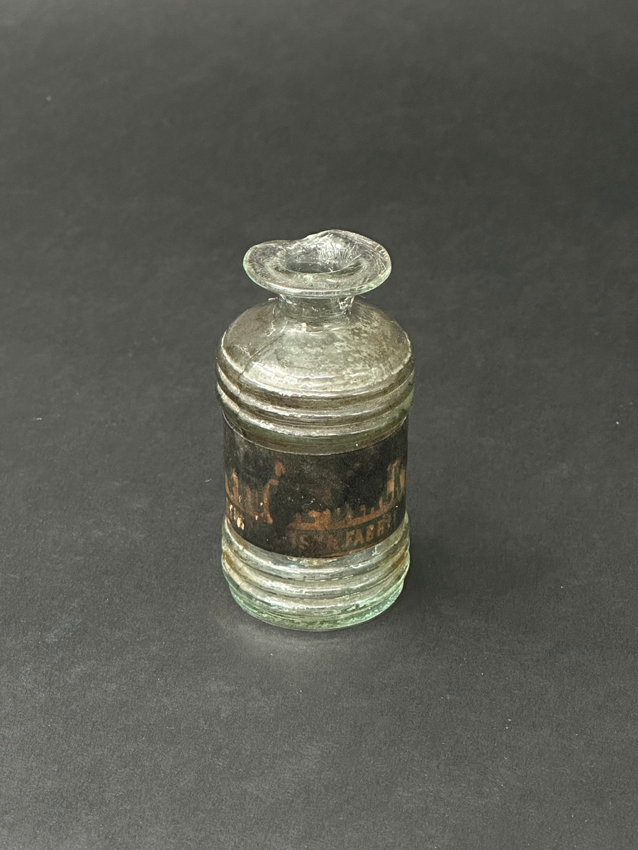 Flaska för bläck. Av glas, ornerad med tre parallella vulster i sin övre och nedre del. Två tydliga lodräta spår i relief efter produktionsform. Tillplattad mynningsrand. En glasflisa har slagits av i mynningen. Mycket ojämn glasmassa med en del lusor. Svart och fläckig etikett som är limmad runt flaskans mitt.