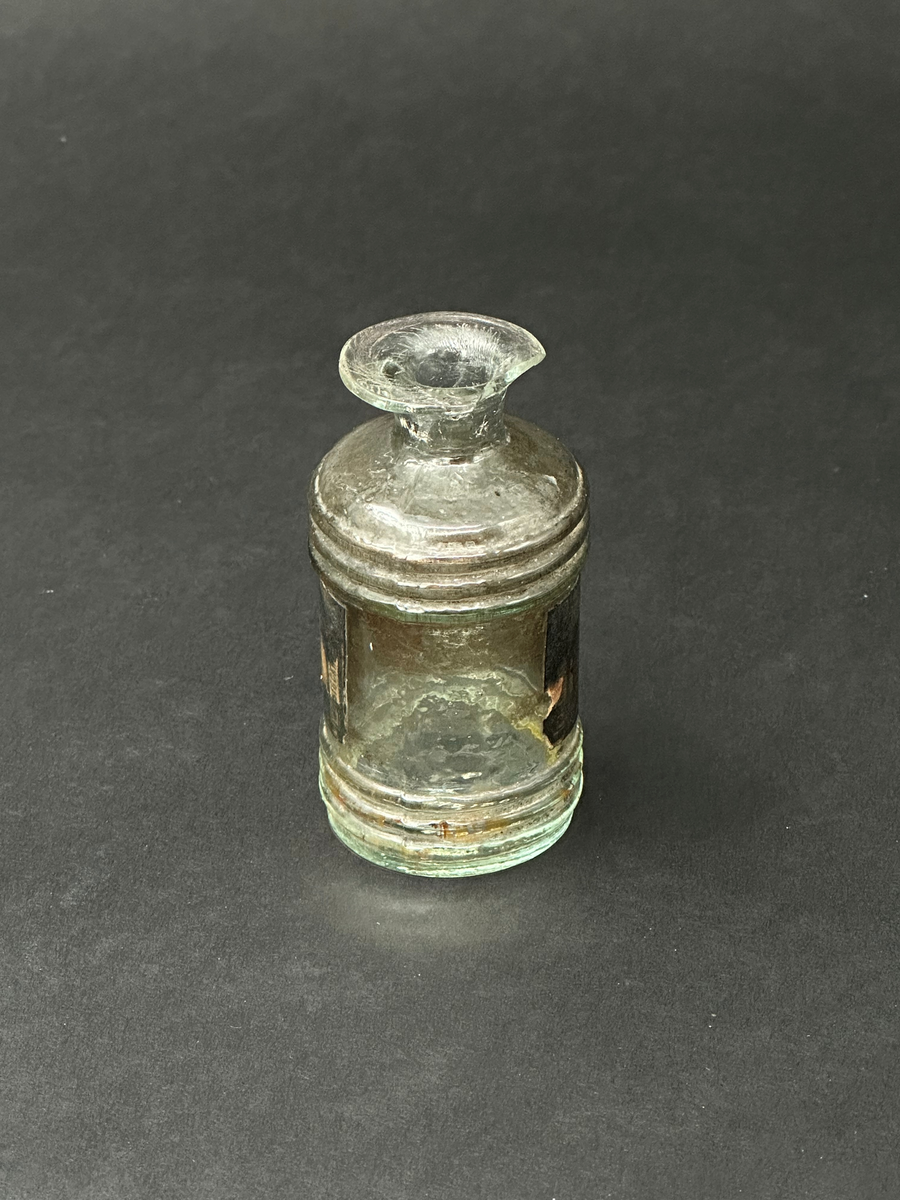 Flaska för bläck. Av glas, ornerad med tre parallella vulster i sin övre och nedre del. Två tydliga lodräta spår i relief efter produktionsform. Tillplattad mynningsrand. En glasflisa har slagits av i mynningen. Mycket ojämn glasmassa med en del lusor. Svart och fläckig etikett som är limmad runt flaskans mitt.