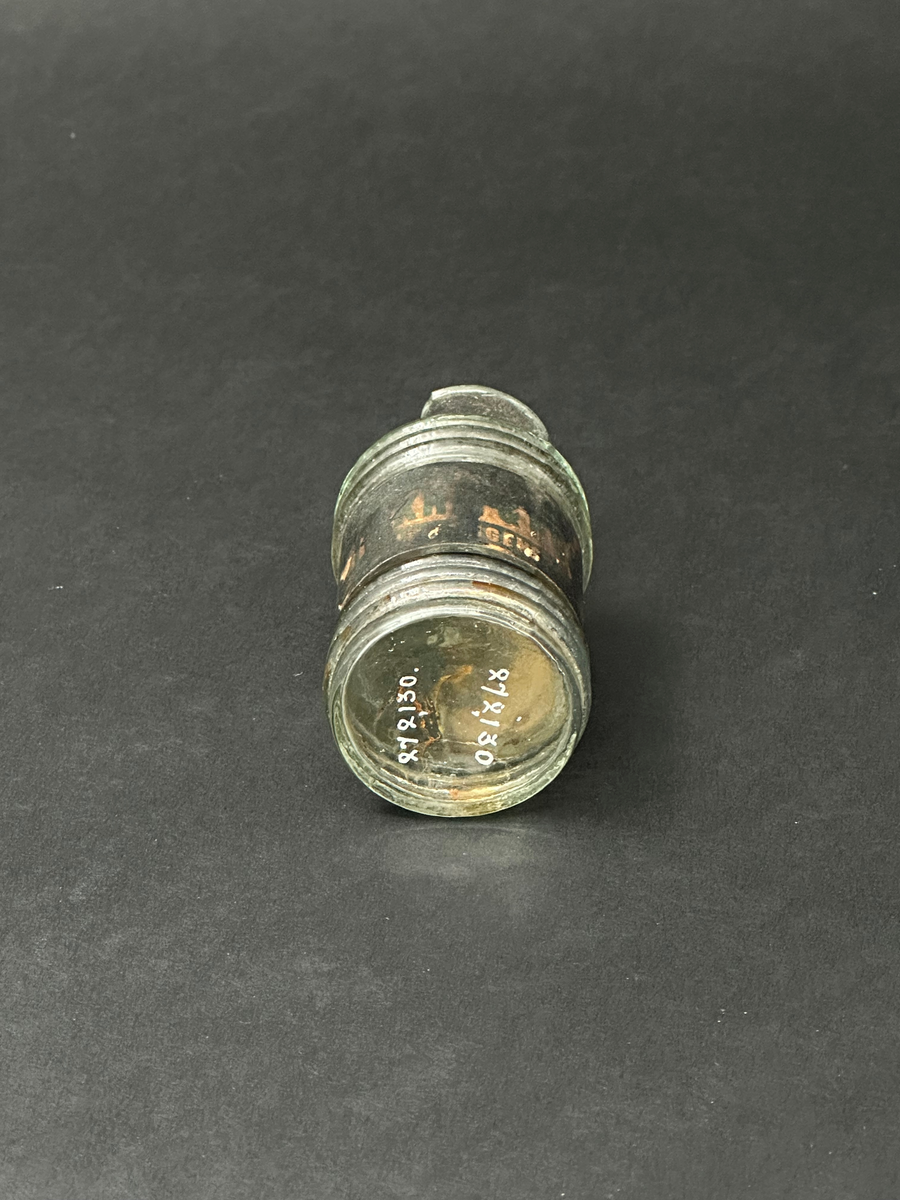 Flaska för bläck. Av glas, ornerad med tre parallella vulster i sin övre och nedre del. Två tydliga lodräta spår i relief efter produktionsform. Tillplattad mynningsrand. En glasflisa har slagits av i mynningen. Mycket ojämn glasmassa med en del lusor. Svart och fläckig etikett som är limmad runt flaskans mitt.