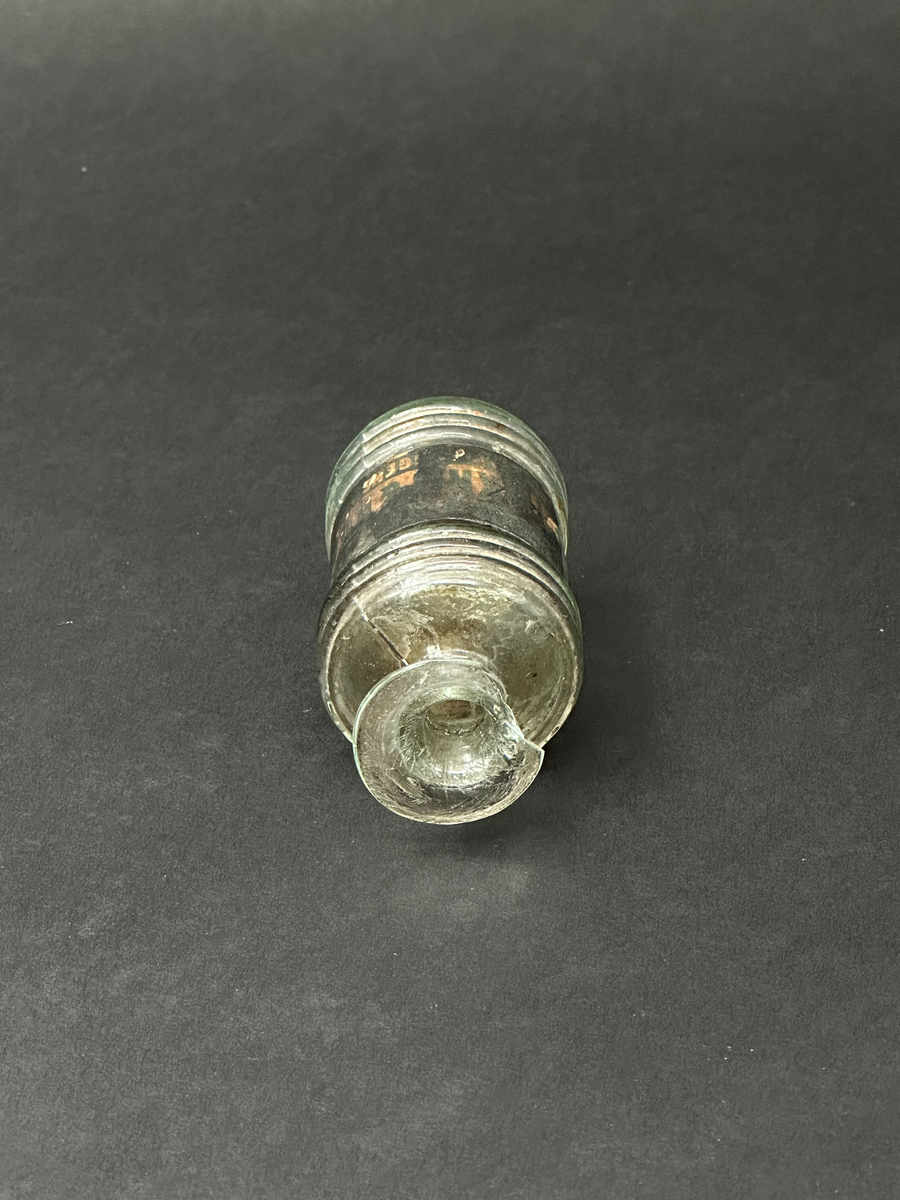 Flaska för bläck. Av glas, ornerad med tre parallella vulster i sin övre och nedre del. Två tydliga lodräta spår i relief efter produktionsform. Tillplattad mynningsrand. En glasflisa har slagits av i mynningen. Mycket ojämn glasmassa med en del lusor. Svart och fläckig etikett som är limmad runt flaskans mitt.