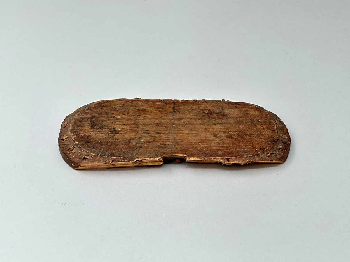 Botten till korg, av trä. Oval. Skuren i ett stycke. C:a 1 cm från ytterkanten vertikala hål som återkommer med 1,5 cm mellanrum. I hålen rester av spröt som syns både från ovan- och nedsidan. Två rektangulära urtag på ömse långsidor av ovalen. Ur ett av urtagen sticker det ut en horisontell tränabb. Ena långsidan av ovalen avknäckt och saknas.