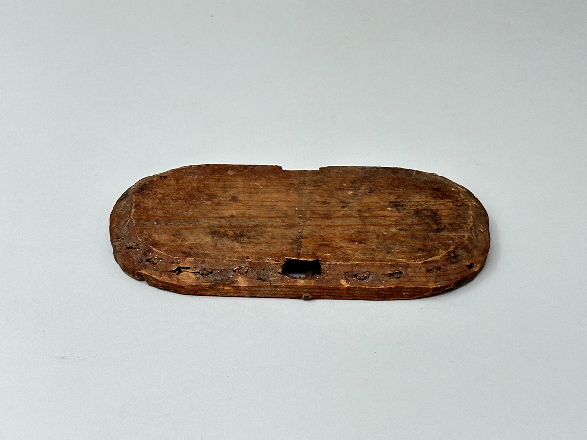 Botten till korg, av trä. Oval. Skuren i ett stycke. C:a 1 cm från ytterkanten vertikala hål som återkommer med 1,5 cm mellanrum. I hålen rester av spröt som syns både från ovan- och nedsidan. Två rektangulära urtag på ömse långsidor av ovalen. Ur ett av urtagen sticker det ut en horisontell tränabb. Ena långsidan av ovalen avknäckt och saknas.