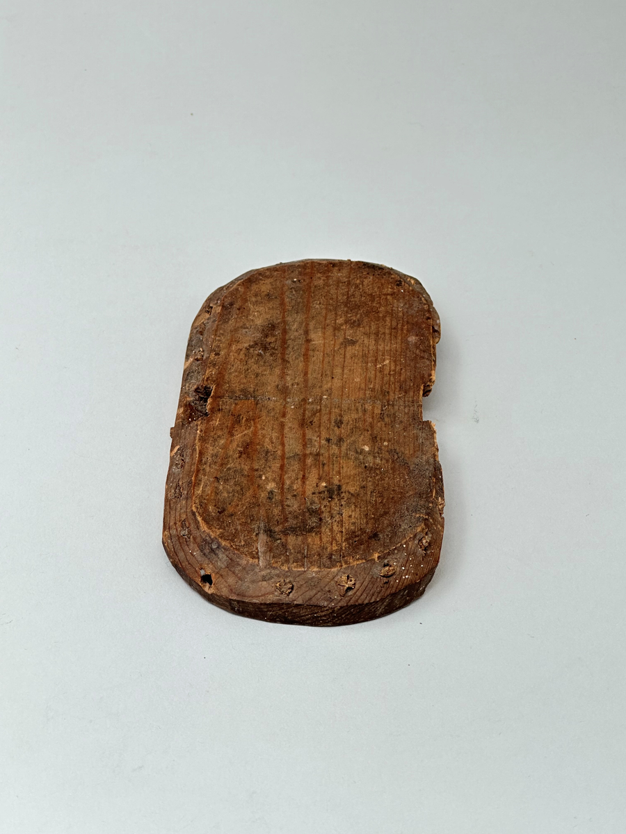 Botten till korg, av trä. Oval. Skuren i ett stycke. C:a 1 cm från ytterkanten vertikala hål som återkommer med 1,5 cm mellanrum. I hålen rester av spröt som syns både från ovan- och nedsidan. Två rektangulära urtag på ömse långsidor av ovalen. Ur ett av urtagen sticker det ut en horisontell tränabb. Ena långsidan av ovalen avknäckt och saknas.