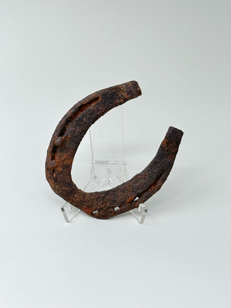 Hästsko i järn. Med fyra hål på vardera sida för söm. Två hål har kvarsittande spik. Kraftigt anfrätt av rost.