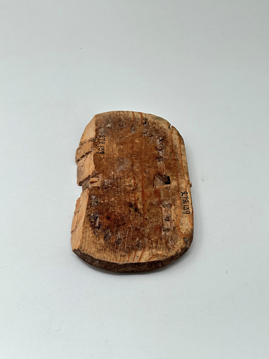 Botten till korg, av trä. Oval. Skuren i ett stycke. C:a 1 cm från ytterkanten vertikala hål som återkommer med 1,5 cm mellanrum. I hålen rester av spröt som syns både från ovan- och nedsidan. Två rektangulära urtag på ömse långsidor av ovalen. Ur ett av urtagen sticker det ut en horisontell tränabb. Ena långsidan av ovalen avknäckt och saknas.